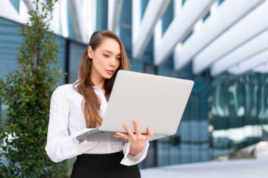 Ofis binasının önünde dizüstü bilgisayar kullanan kendine güvenen bir bayan yönetici. Genç iş kadını beyaz gömlekli. Kadın iş yerinde ağ defteri tutuyor.