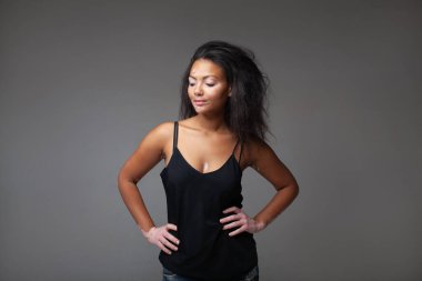 Gri arka planda elleri kalçasında duran duygusal kadın model. Vitiligo cilt hastalığı olan genç bir kadın stüdyoda siyah kolsuz bluz giyiyor.