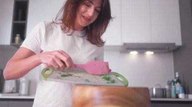 Gündelik kıyafetli gülümseyen bir kadın modern mutfağın pembe spatulasını kullanarak tahtadan yapılmış tahtanın içine doğranmış sebzeler ekliyor ve evde sağlıklı yemek hazırlama sırasında pişirme sürecini gösteriyor.