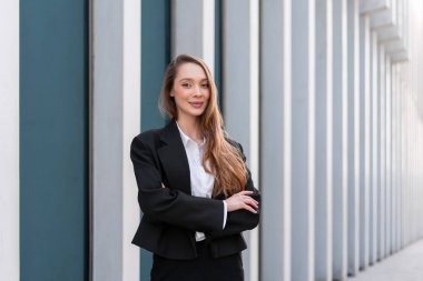 İş elbisesi içinde kollarını kavuşturmuş gülümseyen bir kadının portresi. Şehirdeki bina boyunca kaldırımda uzun saçlı genç bir iş kadını. O, kendinden emin bir şekilde kameraya bakıyor.