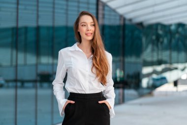 Modern ofis binasının önünde duran, kendine güvenen, uzun saçlı bir iş kadını. Beyaz gömlekli, siyah pantolonlu, elleri ceplerinde, ileriye bakan genç bir kadın.