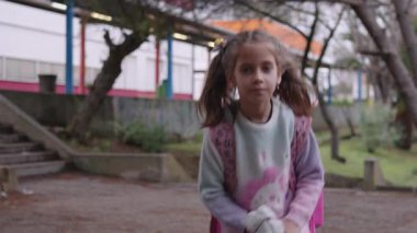 Okul çıkışında at kuyruklu ve pembe sırt çantalı tatlı bir kız öğrenci. Çocuk sakin bir günde kampüsten çıkarken, arka planda ağaçlar ve binalarla mutlu ve rahat görünüyor.