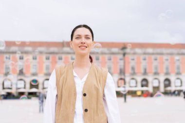Kahverengi yelekli, gülümseyen bir kadın kasaba meydanında kameraya bakıyor. Bulanık binanın önünde poz veren kadın turist, şehir ortamında sıcaklık ve güven yayıyor.