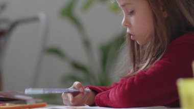 Pijamalı bir kız evdeki mutfakta ödev yapıyor. Kararlı bir ifadeyle öne doğru eğilirken yazmaya odaklanıyor.