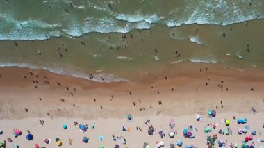 Kumlu kumsalın, renkli şemsiyeli ve güneşli günlerin tadını çıkaran turistli hava aracı görüntüleri. İnsanlar denizde yüzüyor, kıyıda yürüyor ve sıcak kumda dinleniyorlar..