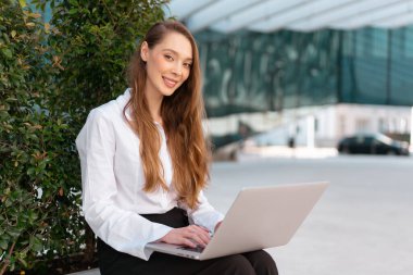 Dışarıda dizüstü bilgisayar kullanan gülümseyen genç iş kadınının portresi. Beyaz gömlekli mutlu genç kadın kameraya bakıyor. Arka planda bulanık ofis binası olan bitkilerin yanında oturuyor.