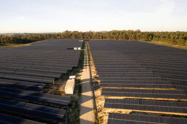 Güneş panelleri sıra sıra uzanan geniş güneş çiftliği manzaraya yayılıyor. Dar toprak yol merkezden geçiyor. Yakınlarda da kamu hizmetleri var. Arka planda sık orman kenarlıkları