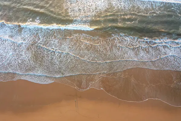 Okyanus dalgalarının kumsala vuruşunu gösteren drone 'lar. Beyaz köpük desenleri, suyun kıyıya çarpmasıyla oluşur. Kum, su kenarında soluk izlerle pürüzsüz görünüyor.