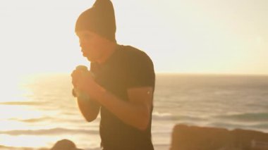 Spor elbiseli sporcu, elinde dambıllarla boks hareketleri yaparken yumruk atıyor. Gün batımında deniz ve dağlar arka planda. Uygun yaşam tarzı ve spor konsepti