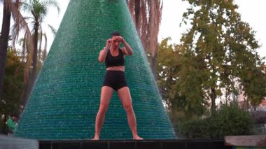 Kadın dövüşçü şehir parkındaki çeşmenin yanında ısınma hareketleri yapıyor. Açık hava kickboks seansından önce odaklanmış siyah spor giyim trenlerinde aktif bir kadın.