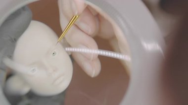 Sanatçıya yakın çekim, büyüteç ve ince fırça kullanarak silikon bebek kafasının üzerine detaylı bir göz çizerek, minyatür sanat sürecinde odak ve hassasiyet gösteriyor.