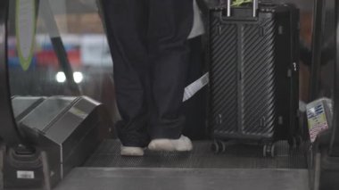 Bavullu gezgin, ulaşım merkezinde yürüyen merdivene biner. Hareketleri, ulaşım rutinini ve kapalı şehir ortamında modern seyahatleri yakalar.