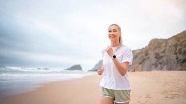 Gülümseyen kadın sabahları kumlu sahilde koşuyor. Beyaz bluzlu atletik kadın ve okyanus kıyısında koşan yeşil şortlu, doğa ve açık havanın tadını çıkaran.