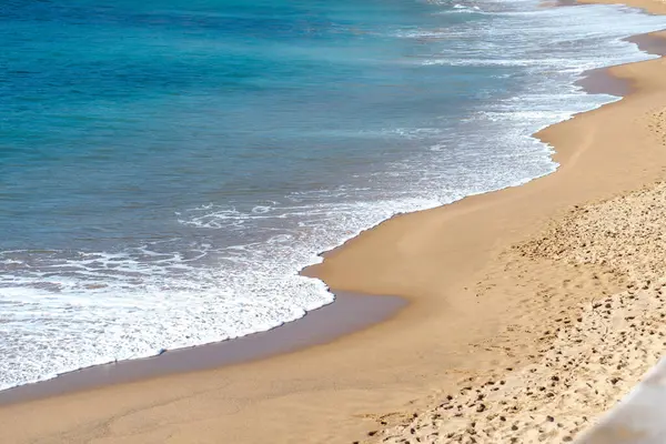 Yumuşak, güzel okyanus dalgaları yavaşça kumlu kumsalda yuvarlanıyor. Turkuaz deniz uzanır, huzurlu ve huzurlu bir kıyı manzarası yaratır. 