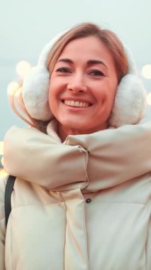 Keyifli bir kadın rahat bir palto giyiyor ve tüylü kulaklıklar kışın ışıl ışıl gülümsüyor. Şenlik ışıklarıyla süslenmiş. Noel zamanı ortam hoş ve neşeli..