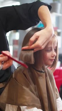 Küçük kız, kuaför koltuğunda güven içinde otururken bir stilist saçını klasik bir Bob 'a çeviriyor. Çocuk, stilisti kusursuz görünümü için makas ve tarak kullanırken gülümsüyor..