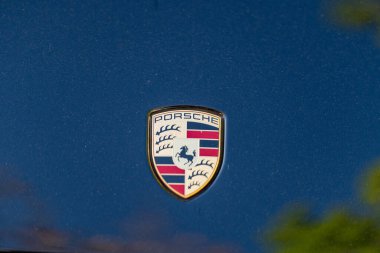 Porsche amblemi, altın rengi ve parlak renkleri bulunan bir araçta belirgin bir şekilde sergilenmektedir. Yansımalar ve mavi bir arkaplan zarafeti ve lüksü artırır..