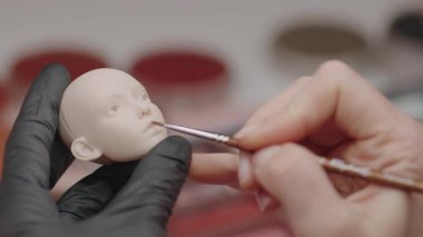 Sanatçının eldiven giyerken ince fırça kullanarak bebek kafasını boyaması, hassas özelliklere odaklanması ve yaratıcı çalışmalar sırasında ince detaylar üzerinde durması.