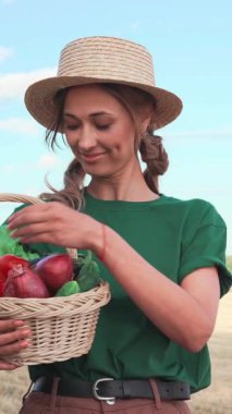 Kadın açık havada elinde renkli sebzelerle dolu bir sepet tutarken gülümsüyor. Etrafı doğa ve taze ürünlerle çevrili, sağlıklı bir yaşam tarzını benimseyerek zaman geçirmekten hoşlanıyor..
