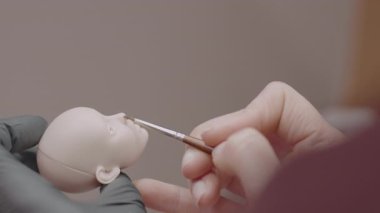 Eldivenli elle tutulan silikon bebek kafasını boyamak için ince fırça kullanan işçilere yakın çekim, yüz detaylarına odaklanma ve dikkatli el işi sanatları.