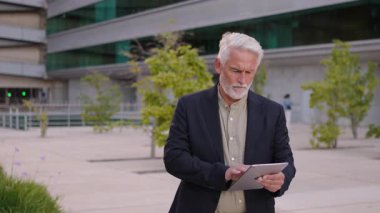 Şehir merkezindeki açık alanda tablet kullanan gri saçlı, kendinden emin kıdemli bir işadamı. Şık bir kıyafet giyiyor ve şehir ticaret bölgesine odaklanmış gibi görünüyor.