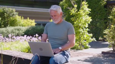 Parktaki bankta otururken dizüstü bilgisayarda çalışan kıdemli iş adamı. Şık giyinmiş, gün ışığına odaklanmış görünüyor. Etrafında yeşillik var.