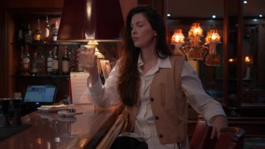 Barda oturmuş viski içen, düşünceli bir ifadeyle gözlerini kaçıran bir kadın. Sıcak ışıklarla şık bir ortam, sakin ve şık bir ortam yaratır. Yansımanın sessiz anlarını yakalar.