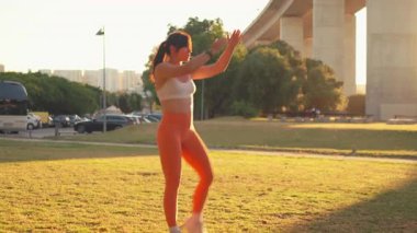 Arka planda günbatımı ile yüksek diz egzersizi yapan bir kadın. Beyaz hasat bluzu ve turuncu tozluklarıyla antrenman sırasında park yerinde formda kalıyor.