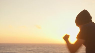 Kararlı sporcu adam gün batımında dağlarla ve gökyüzüyle deniz manzarasına bakarken hava boksu yapıyor. Güçlü, zinde bir genç adam doğada ara sıra eğitim yapıyor.