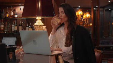 Şık bir barda durup viski içen bir iş kadını. Tezgahta dizüstü bilgisayar, sıcak aydınlatma ve zarif atmosfer, rahat çalışma öneriyor.