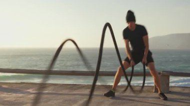 Atletik adam gün batımında deniz kenarındaki savaş halatlarıyla antrenman yapıyor. Spor, spor ve sağlıklı yaşam tarzı konsepti. Kaslı vücutlu beyaz sporcu fonksiyonel eğitim ve dayanıklılığa odaklandı 
