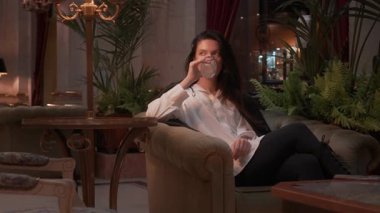 İş kadını resmi kıyafetiyle camdan viski içerken aynı zamanda şık bir salonda sıcak ışıklandırma, iç mekan bitkileri ve arka planda görünen lüks iç dekorları olan bir kanepede oturuyor. 