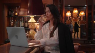 Beyaz gömlekli bir iş kadını barda oturmuş viski içiyor ve dizüstü bilgisayarla çalışıyor. Sıcak ve sofistike ortam iş ve rahatlama dengesini vurguluyor.