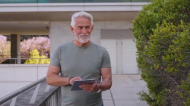 Modern binanın yanında tablet kullanırken beyaz saçlı ve sakallı olgun bir adam yürüyor. Odaklanmış ve düşünceli görünüyor.