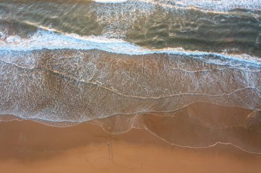 Yukarıdan aşağıya deniz dalgalarının boş sahildeki ıslak kumun üzerinde yavaşça yuvarlanan drone görüntüsü. Sudaki doğal desenler kıyı şeridindeki sakin ve soyut kıyı sahnesini oluşturur.