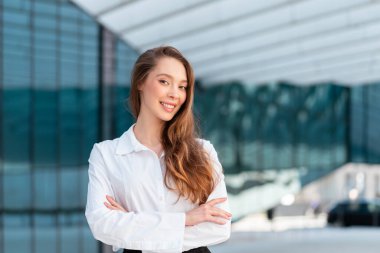 Beyaz gömlekli kendine güvenen iş kadını modern cam ofis yakınlarında kollarını kavuşturup gülümsüyor, başarısını, liderliğini ve profesyonel varlığını gösteriyor.