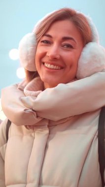 Neşeli kadın sıcak bir palto ve kulaklık giyerken gülümsüyor. Canlı kış ayinlerinde sıcak ışıklar, tatil sezonunun ruhunu yakalama ve neşeli tezahüratlar yer alıyor..