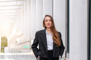 Siyah takım elbiseli ve beyaz gömlekli iş kadını modern şehir bölgesinde kendine güvenen bir gülümseme ile ayakta duruyor. Uzun saçlı genç bir profesyonel, güpegündüz ofis binalarının yakınındaki kameraya bakıyor.