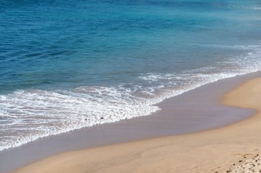Yumuşak, güzel okyanus dalgaları yavaşça kumlu kumsalda yuvarlanıyor. Turkuaz deniz uzanır, huzurlu ve huzurlu bir kıyı manzarası yaratır. 