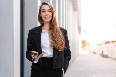 Resmi siyah takım elbiseli, beyaz gömlekli, akıllı telefon tutan, modern ofisin önünde gülümseyen, başarılı ve profesyonelce davranan kendine güvenen bir kadın.