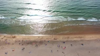Sahilde dalgalar kıyıya vururken dinlenen insanlar. Köpüklü su ve altın kum mükemmel plaj günü için sakin bir atmosfer yaratır.