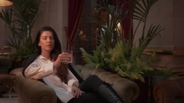 Resmi kıyafetli bir iş kadını, camdan viski içerken aynı zamanda şık bir salonda, arka planda sıcak ışıklandırma, iç mekan bitkileri ve lüks iç dekorları olan bir kanepede oturuyor.