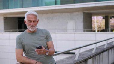 Senior businessman with white hair and beard confidently holding smartphone in hand engaging in conversation with positive expression. Male professional leaning on railing outside workspace