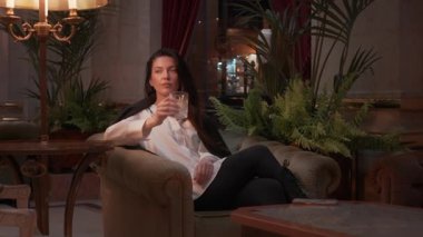Woman in formal outfit drinking whiskey from glass while sitting on couch in elegant lounge with warm lighting, indoor plants and luxurious interior decor visible in background 