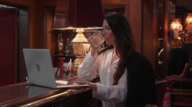Şık bir barda durup viski içen bir iş kadını. Tezgahta dizüstü bilgisayar, sıcak aydınlatma ve zarif atmosfer, rahat çalışma öneriyor.