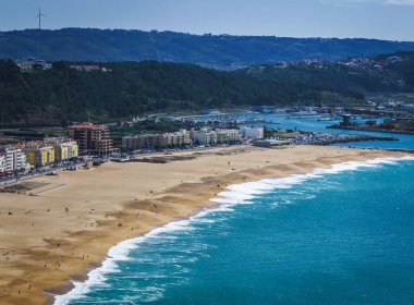 Yukarıdan okyanus kıyısının manzarası, kumlu sahil boyunca mavi-yeşil suları ve beyaz dalgaları gösteriyor. Portekiz 'in Nazare kenti yakınlardadır. Güneşli bir gün.