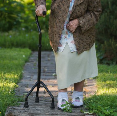 Bastonlu yaşlı bir kadın kırsalda asfalt bir yol boyunca yürür, bir bahçede; vücudun görünür bir parçası.