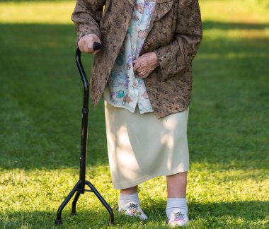 Bastonlu yaşlı bir kadın yeşil çimlerin üzerinde yürüyor kırsalda, bahçede; vücudun görünür bir parçası.