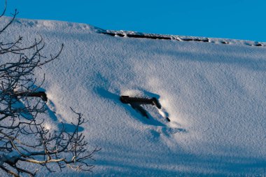 Evin çatısı ışıltılı bir kar tabakasıyla kaplı, karın altında iki pencere; çatının arkasında açık mavi bir gökyüzü var..