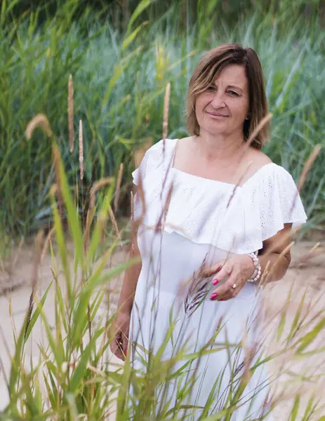 Yazın deniz kenarında, beyaz elbiseli bir kadın bize gülümserken bakar, bir kum tepeciğindeki çimlerin arkasında durur..